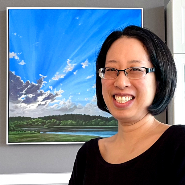 Photo of a smiling Asian woman with chin length hair and glasses. She is in front of a painting of a blue sky with the sun shining from behind some clouds.